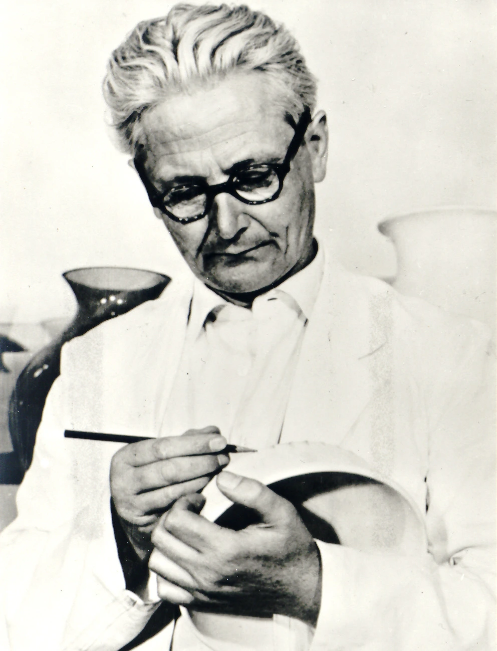 Wilhelm Wagenfeld wearing glasses, writing on a white bowl with a black pencil