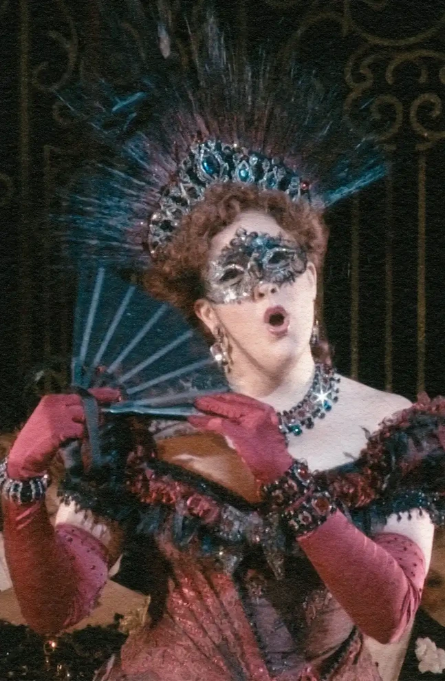 A performer in an elaborate red Victorian ballroom gown with silk, ruffles, rhinestones, and feathers, holding a black fan and mask for the MET Opera production of 'Die Fledermaus,' supported by Bloomberg Philanthropies.