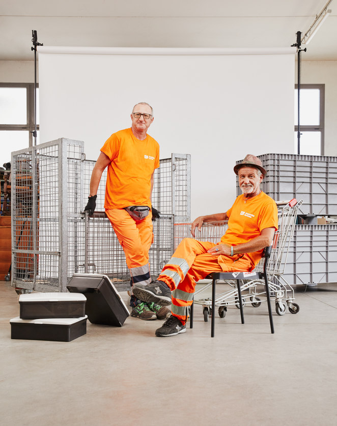Fotografia di due dipendenti che lavorano all'ecocentro. Con nello sfondo dei carrelli e delle grate che richiamano l'ambiente dell'ecocentro