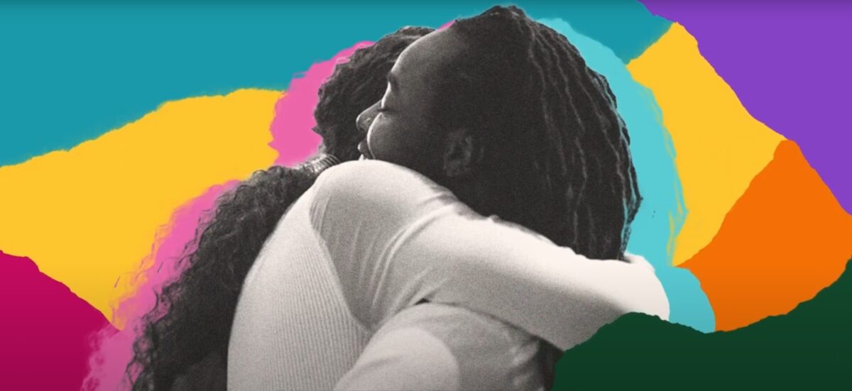 Two Black, trans figures embrace, with a collage of brightly colored paper scraps as their background.