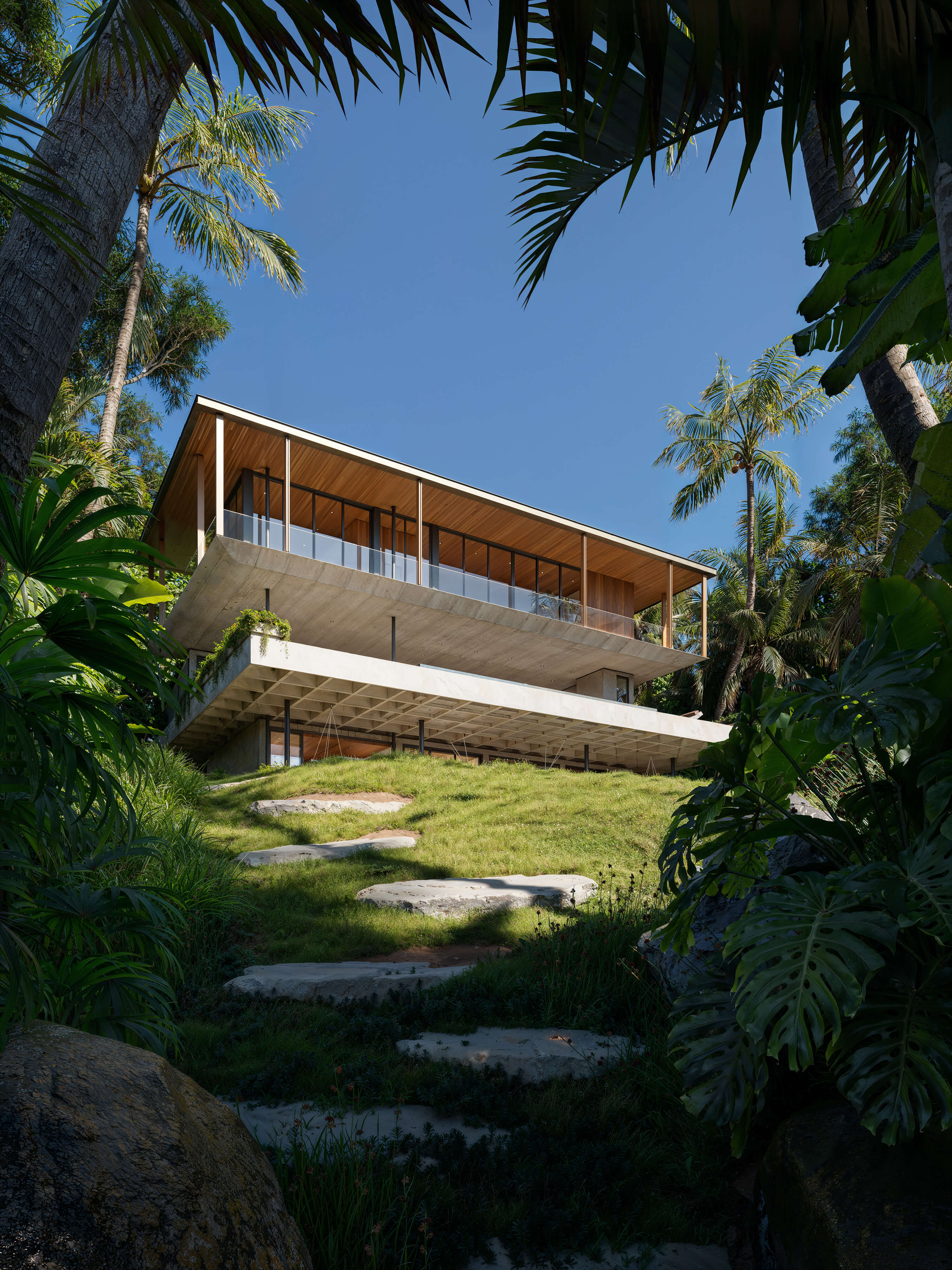 Tropical hillside CGI of a modern concrete and wood residence with cantilevered upper floor, full-height glass walls, and lush vegetation; architectural visualization emphasizing elevation and natural integration.

