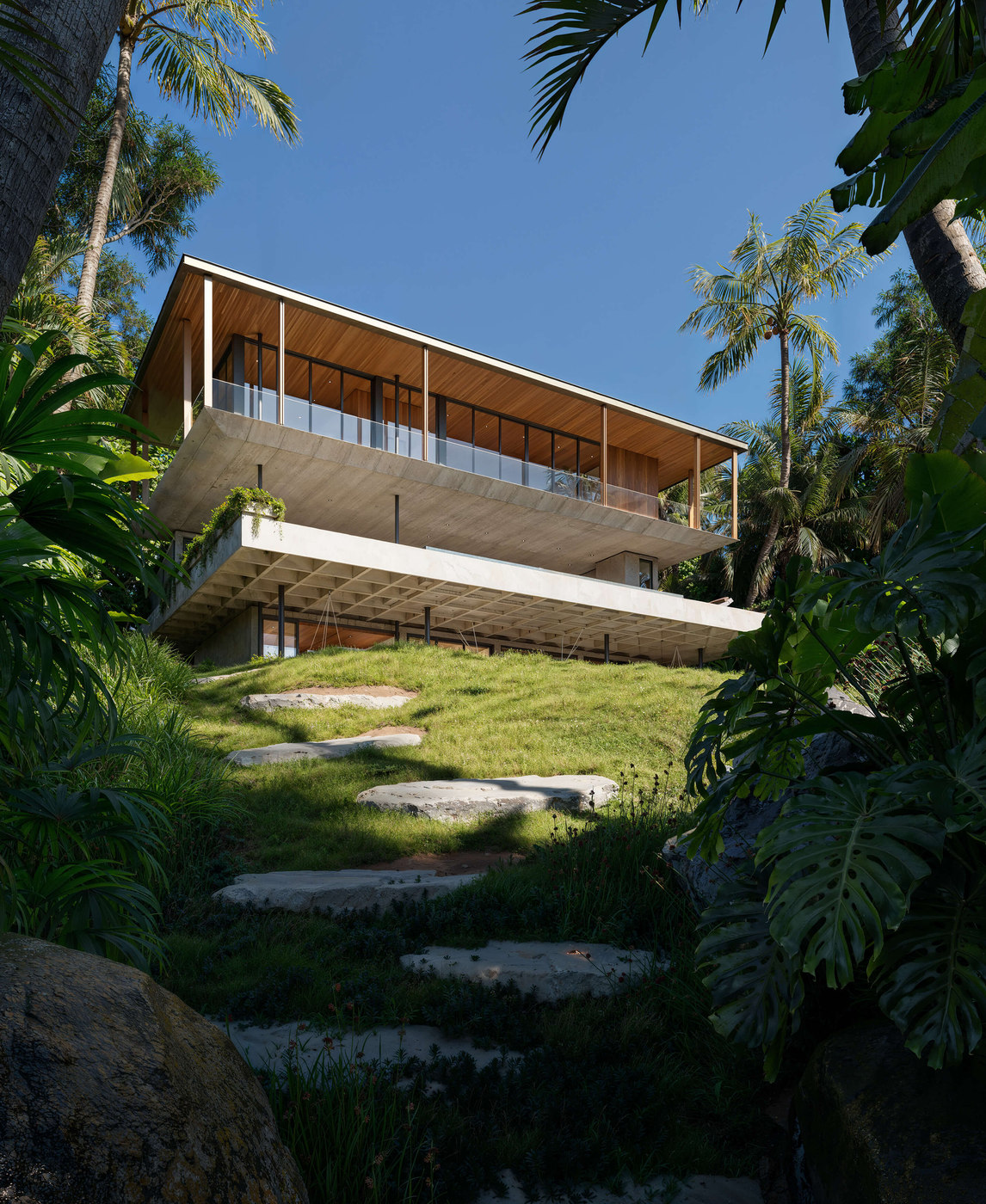 Tropical hillside CGI of a modern concrete and wood residence with cantilevered upper floor, full-height glass walls, and lush vegetation; architectural visualization emphasizing elevation and natural integration.