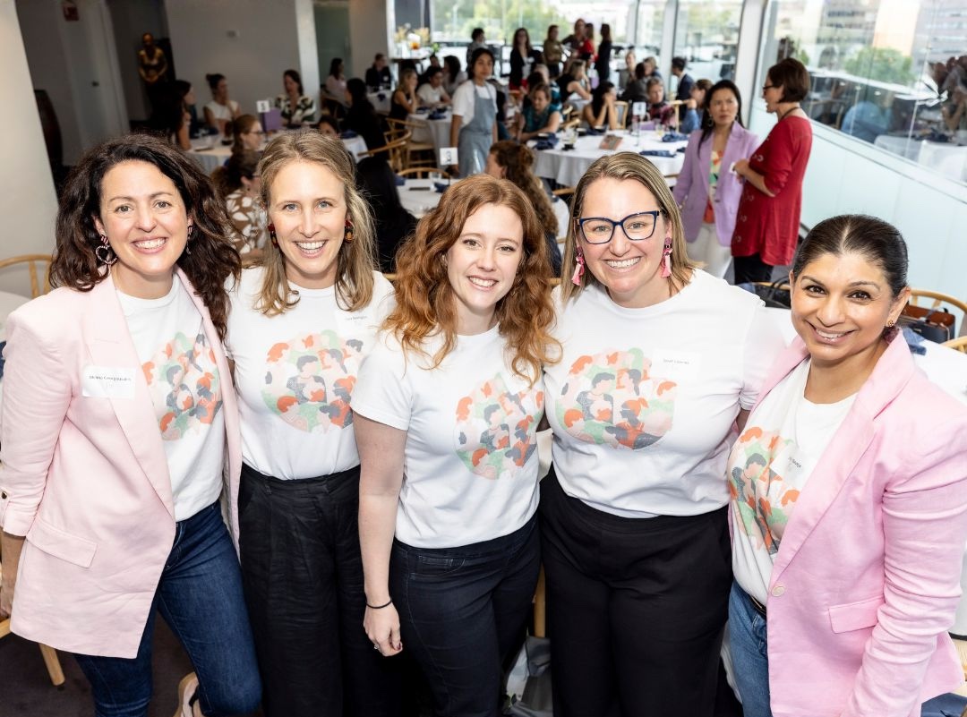 The Franklin Women team standing together and smiling at a community event, wearing Franklin Women T-shirts, with attendees and tables visible in the background.
