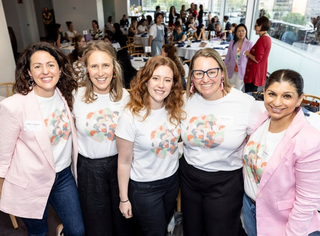 The Franklin Women team standing together and smiling at a community event, wearing Franklin Women T-shirts, with attendees and tables visible in the background.