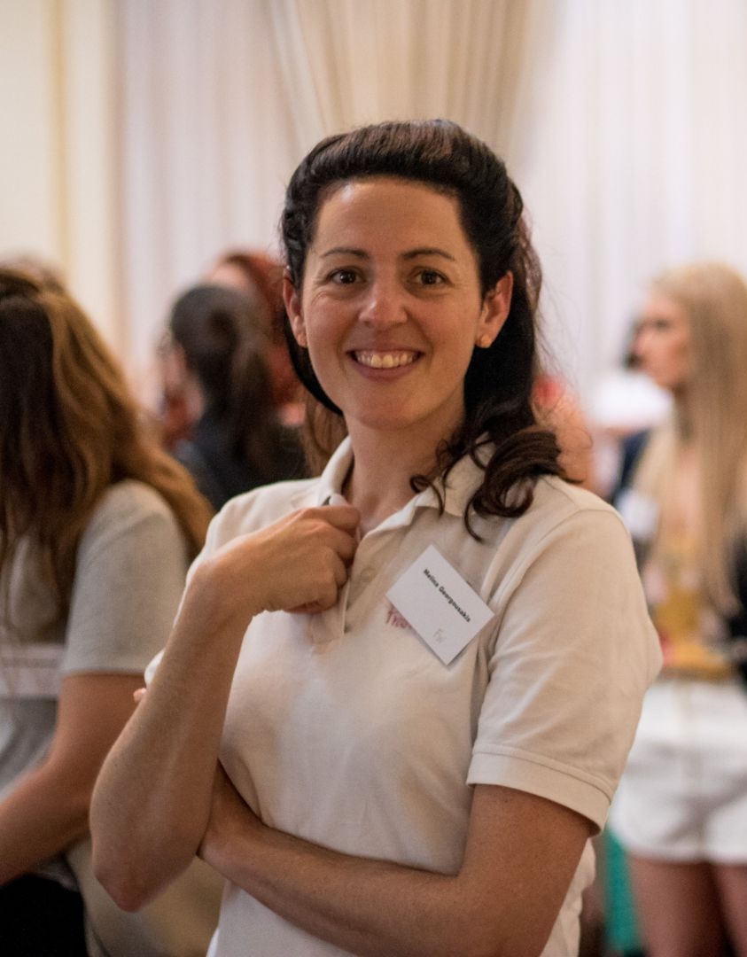 Dr Melina Georgousakis smiling at a Franklin Women event, wearing a white polo shirt and name badge, standing with arms crossed in a room of attendees.