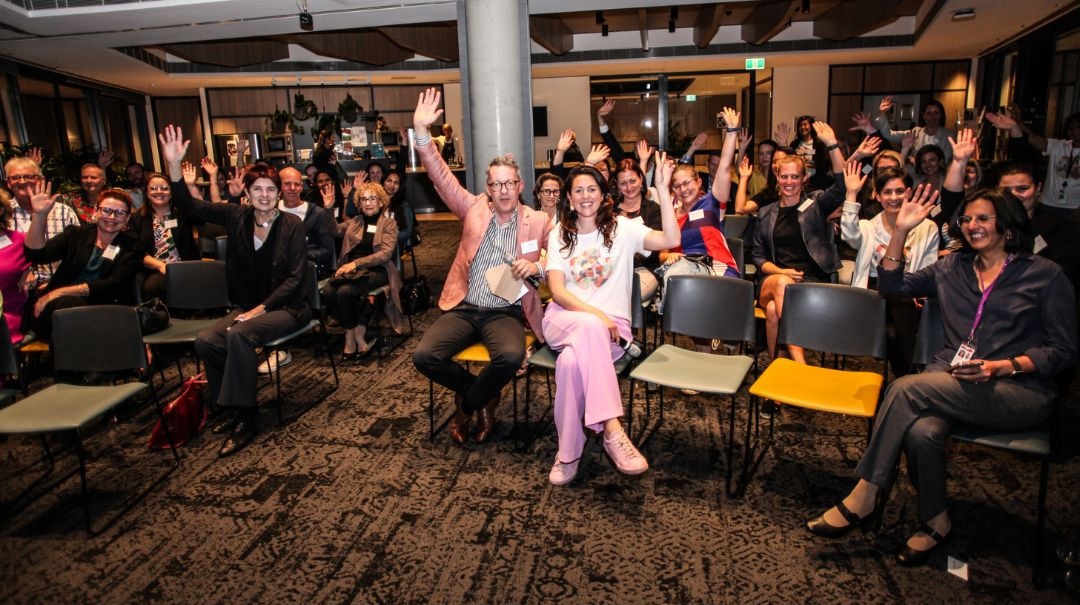Attendees at a Franklin Women event seated in a room, smiling and raising their hands toward the camera.