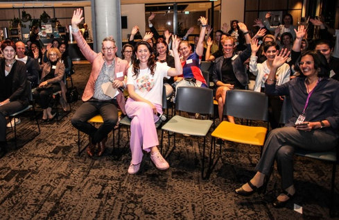 Attendees at a Franklin Women event seated in a room, smiling and raising their hands toward the camera.